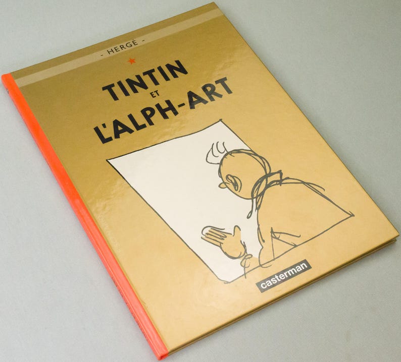 Może przedstawiać: Twarda oprawa książki "Tintin et l'Alph-Art" autorstwa Herg&eacute;. Złota okładka z czarnym tytułem i szkicem Tintina. Grzbiet w jaskrawym pomarańczu. Wydane przez Casterman.