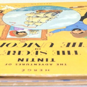 May include: A vintage hardcover book titled "The Secret of the Unicorn" from "The Adventures of Tintin" series. The cover is yellow with illustrations and text. The book's spine is red.