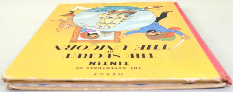 May include: A vintage hardcover book titled "The Secret of the Unicorn" from "The Adventures of Tintin" series. The cover is yellow with illustrations and text. The book's spine is red.