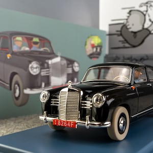 Puede incluir: Un modelo de coche Mercedes-Benz negro con neum&aacute;ticos de banda blanca y una matr&iacute;cula roja. El modelo est&aacute; sobre una base azul, con una caja que presenta una ilustraci&oacute;n de coche similar en el fondo. La escala es 1:24.