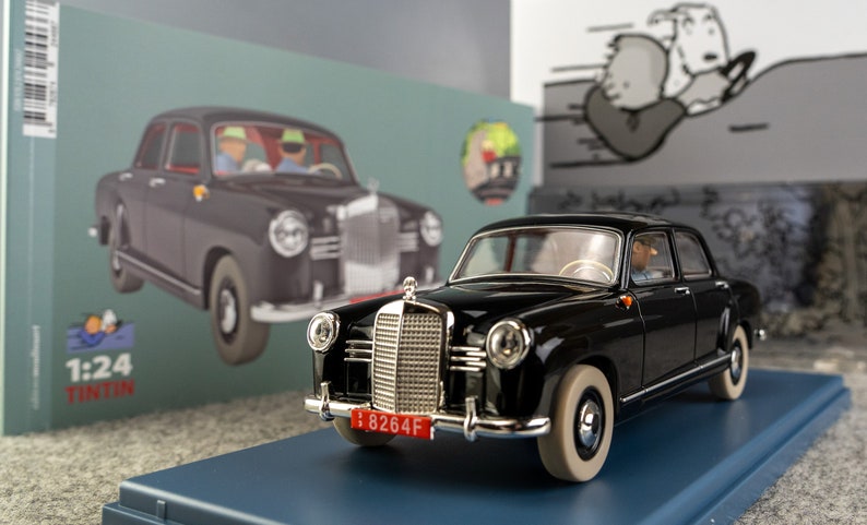 Puede incluir: Un modelo de coche Mercedes-Benz negro con neum&aacute;ticos de banda blanca y una matr&iacute;cula roja. El modelo est&aacute; sobre una base azul, con una caja que presenta una ilustraci&oacute;n de coche similar en el fondo. La escala es 1:24.