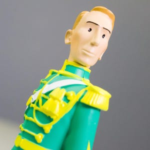 May include: A figurine of a man in a green military-style jacket with yellow accents and epaulettes. The figure has blonde hair and is looking to the side. A white strap crosses the chest. The background is a solid gray.