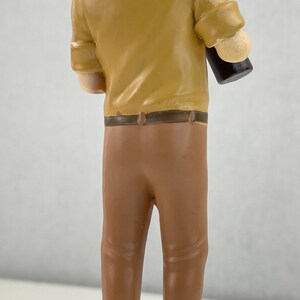 May include: A figurine of a man wearing a white sailor hat and a brown shirt and pants. He is holding a brown bottle in his right hand.