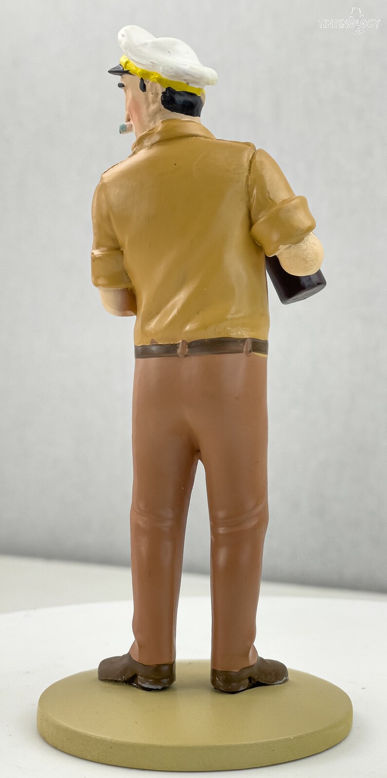 May include: A figurine of a man wearing a white sailor hat and a brown shirt and pants. He is holding a brown bottle in his right hand.