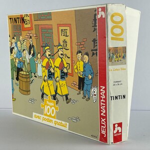 Peut inclure: Un puzzle de 100 pi&egrave;ces repr&eacute;sentant une sc&egrave;ne de la bande dessin&eacute;e de Tintin "Le Lotus Bleu". La bo&icirc;te du puzzle est rouge, blanche et bleue avec le titre "Tintin" et "Le Lotus Bleu" imprim&eacute; dessus. Le puzzle est fabriqu&eacute; par Jeux Nathan.