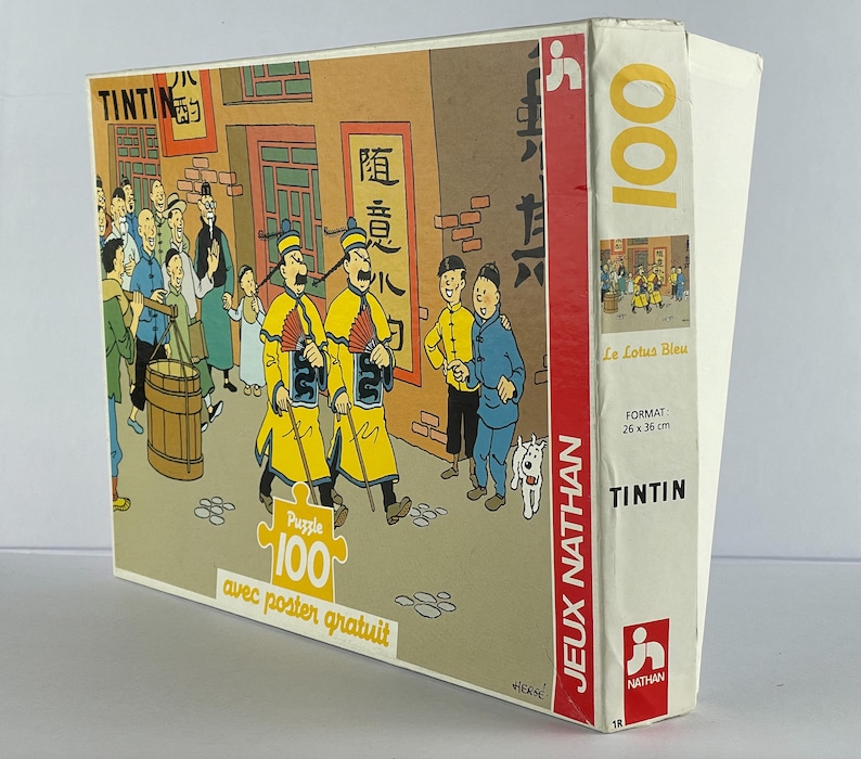 Peut inclure: Un puzzle de 100 pi&egrave;ces repr&eacute;sentant une sc&egrave;ne de la bande dessin&eacute;e de Tintin "Le Lotus Bleu". La bo&icirc;te du puzzle est rouge, blanche et bleue avec le titre "Tintin" et "Le Lotus Bleu" imprim&eacute; dessus. Le puzzle est fabriqu&eacute; par Jeux Nathan.