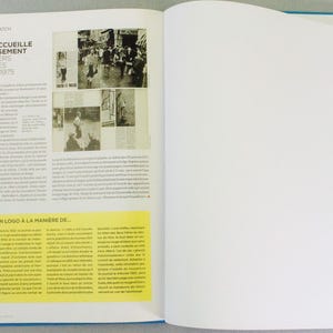 May include: An open book with a blue cover, featuring text and images. The left page has text and black and white photos, with a yellow box containing text and logos. The right page is blank. The text includes "HADDOCK ACCUEILLE CHALEUREUSEMENT LES REPORTERS AU DEBUT DES PICAROS EN 1975".