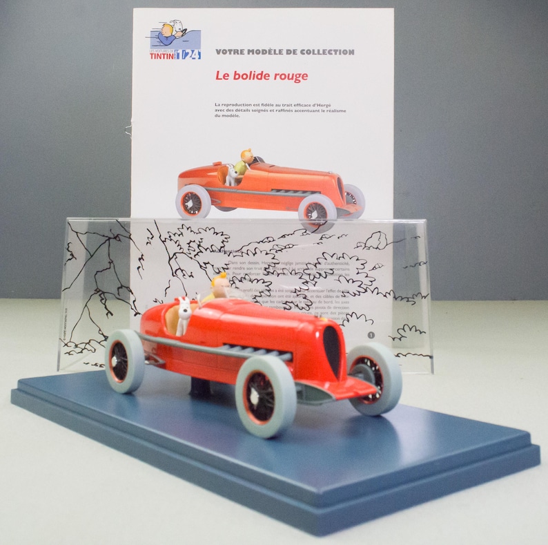 Pu&ograve; includere: Modello di auto da corsa giocattolo rosso con figura di pilota, esposto su una base blu. Il modello &egrave; una replica di "Le bolide rouge" ed &egrave; accompagnato da una scheda stampata con testo francese.