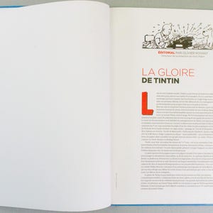 May include: Open book with a blue border. The right page features text in French with the title "LA GLOIRE DE TINTIN" in red. An illustration of a man typing on a typewriter with a dog is at the top.