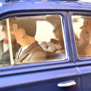 May include: A blue toy car with figures inside. The figures are wearing suits and hats. The car's windows are clear, and the interior is visible. The car is a classic style.
