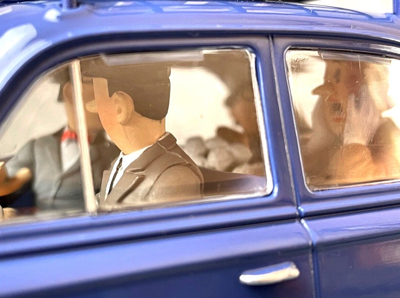 May include: A blue toy car with figures inside. The figures are wearing suits and hats. The car's windows are clear, and the interior is visible. The car is a classic style.