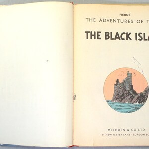K&ouml;nnte beinhalten: Aufgeschlagenes Buch mit dem Titel "Die Schwarze Insel" von Herg&eacute;. Das Cover zeigt eine runde Illustration einer felsigen Insel mit Burg und Meer. Der Verlag des Buches ist Methuen & Co Ltd.