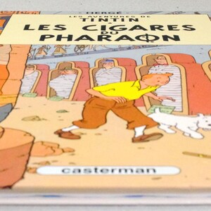 Pode incluir: Um livro de capa dura intitulado "LES CIGARES DU PHARAON" de Herg&eacute;. A capa apresenta uma ilustra&ccedil;&atilde;o de desenho animado de um homem com uma camisa amarela e cal&ccedil;as marrons, correndo com um cachorro branco. O fundo retrata imagens com tema eg&iacute;pcio.