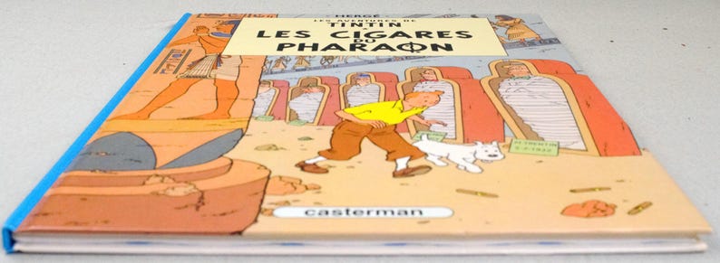 Pode incluir: Um livro de capa dura intitulado "LES CIGARES DU PHARAON" de Herg&eacute;. A capa apresenta uma ilustra&ccedil;&atilde;o de desenho animado de um homem com uma camisa amarela e cal&ccedil;as marrons, correndo com um cachorro branco. O fundo retrata imagens com tema eg&iacute;pcio.