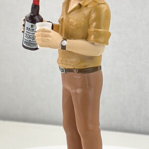 May include: A figurine of a man wearing a brown uniform and a white sailor hat with a gold anchor. He is holding a bottle of Haig's Gold Label Scotch Whisky and a glass.