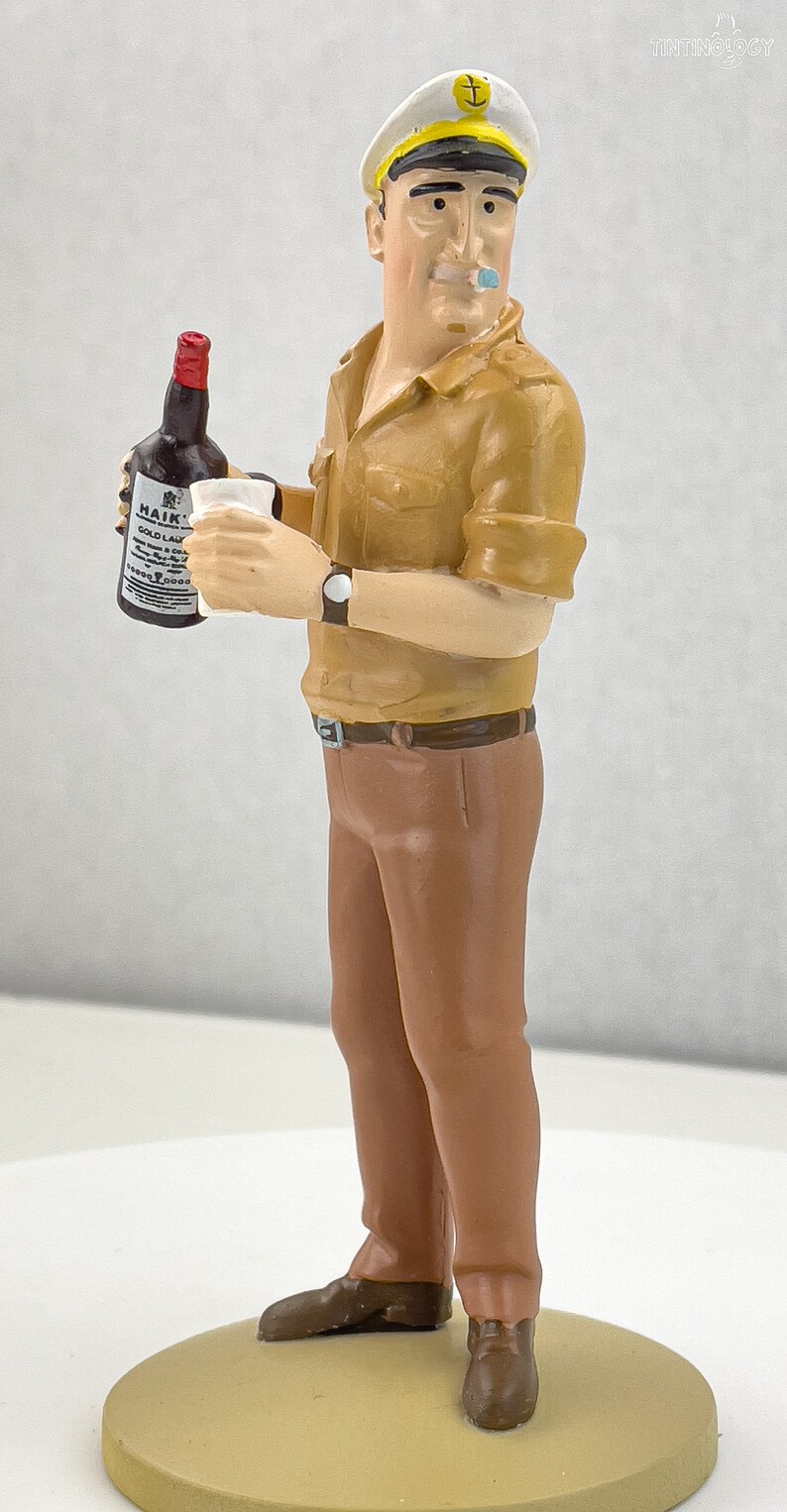May include: A figurine of a man wearing a brown uniform and a white sailor hat with a gold anchor. He is holding a bottle of Haig's Gold Label Scotch Whisky and a glass.