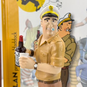 May include: A figurine of a cartoon character wearing a white sailor hat with a gold anchor and a brown shirt. The character is holding a bottle of liquor and a glass. The background is a blurred image of a comic book.