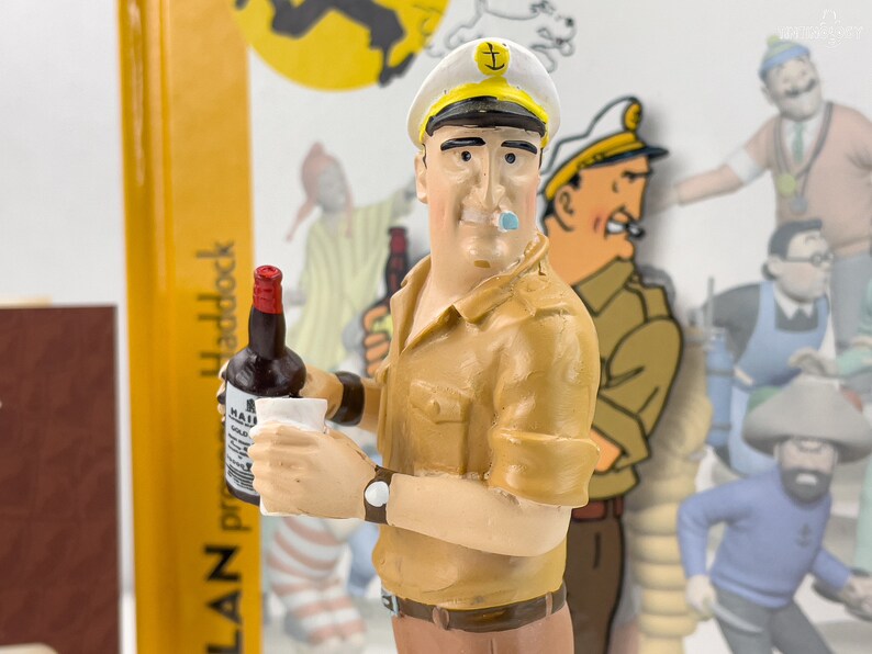 May include: A figurine of a cartoon character wearing a white sailor hat with a gold anchor and a brown shirt. The character is holding a bottle of liquor and a glass. The background is a blurred image of a comic book.