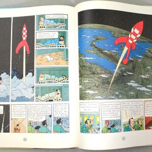 K&ouml;nnte beinhalten: Offenes Buch mit Comic-Strip-Illustrationen. Eine rot-wei&szlig;e Rakete startet ins All, mit Blick auf die Erde im Hintergrund. Weitere Panels zeigen Szenen mit Charakteren in Betten und einem Kontrollraum.