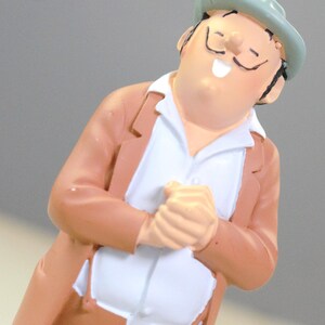 May include: A small figurine of a person with a mustache wearing a light green hat, a tan jacket, and a white shirt. The figure has a joyful expression with eyes closed and hands clasped together in front of the chest, rendered in a smooth finish.