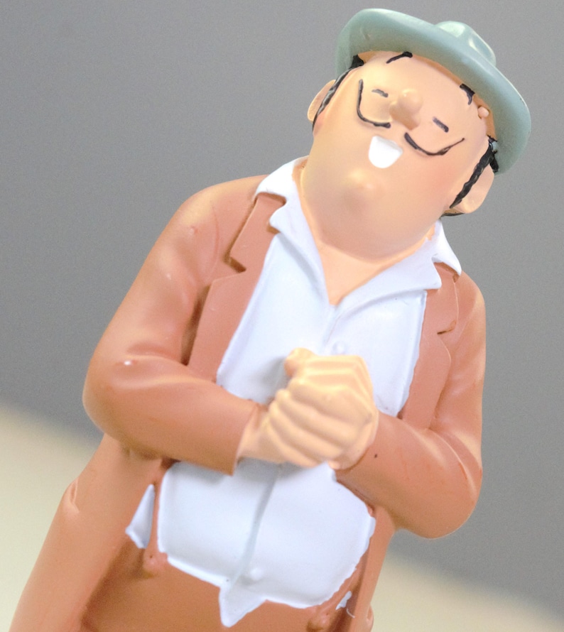 May include: A small figurine of a person with a mustache wearing a light green hat, a tan jacket, and a white shirt. The figure has a joyful expression with eyes closed and hands clasped together in front of the chest, rendered in a smooth finish.