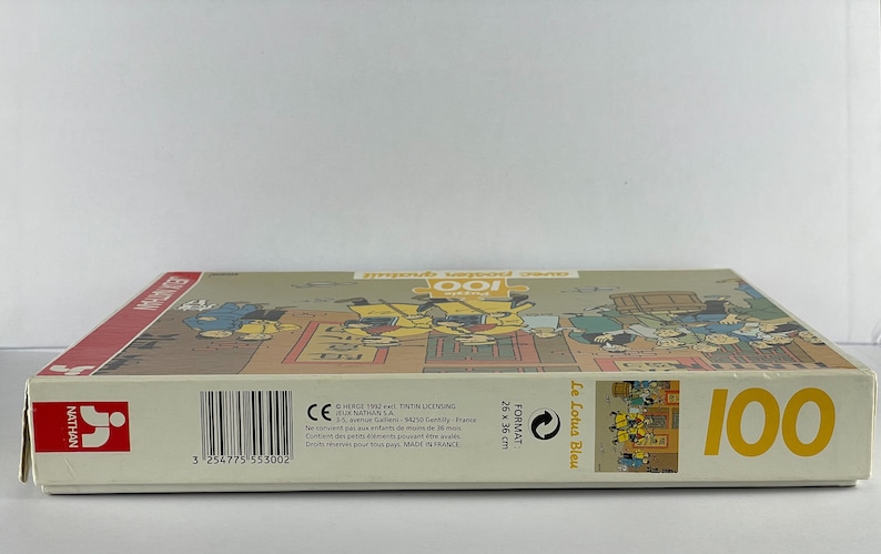 Peut inclure: Bo&icirc;te de puzzle de 100 pi&egrave;ces illustrant des personnages de la bande dessin&eacute;e "Le Lotus Bleu" d'Herg&eacute;. La bo&icirc;te est blanche avec des accents jaunes et le texte "Le Lotus Bleu" et "100" imprim&eacute; dessus. Le puzzle est fabriqu&eacute; en France.