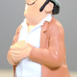 May include: A figurine of a character wearing a light blue fedora with a black band, a tan jacket, and a white collared shirt. The character has dark hair, a thin mustache, and is depicted with hands clasped together in front of the chest.
