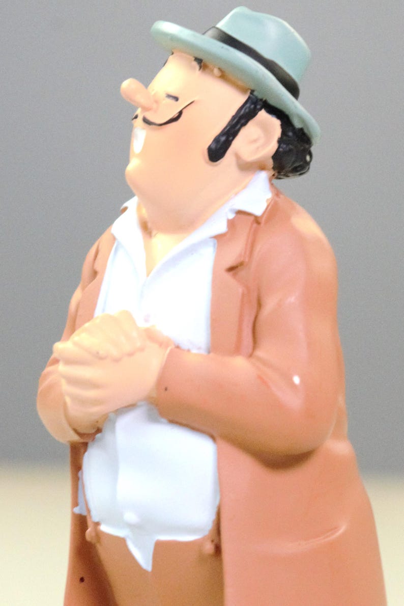 May include: A figurine of a character wearing a light blue fedora with a black band, a tan jacket, and a white collared shirt. The character has dark hair, a thin mustache, and is depicted with hands clasped together in front of the chest.