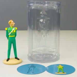 May include: A figurine of a man in a green uniform with gold accents, standing on a yellow base. A clear plastic container holds a similar figure. Two blue oval stickers with matching illustrations are also present.