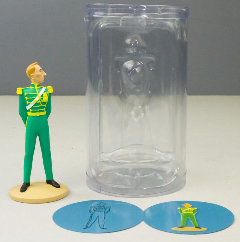 May include: A figurine of a man in a green uniform with gold accents, standing on a yellow base. A clear plastic container holds a similar figure. Two blue oval stickers with matching illustrations are also present.