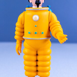May include: A yellow astronaut figurine with a bald head, black mustache, and glasses. The figurine is wearing a yellow spacesuit with blue accents and is standing on a tan base.