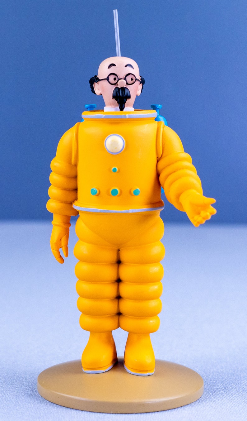 May include: A yellow astronaut figurine with a bald head, black mustache, and glasses. The figurine is wearing a yellow spacesuit with blue accents and is standing on a tan base.