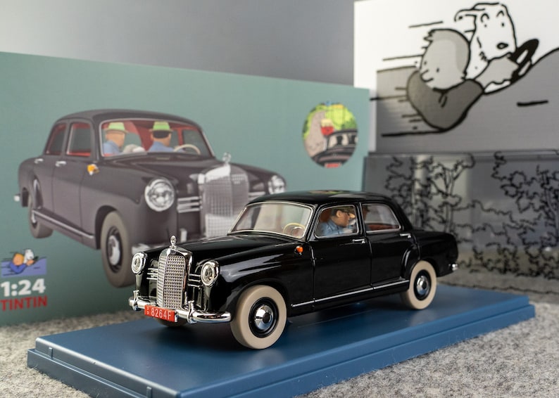 Puede incluir: Un modelo de coche Mercedes-Benz negro con neum&aacute;ticos blancos, expuesto sobre una base azul. El modelo presenta una parrilla cromada detallada y una matr&iacute;cula roja. La caja muestra un coche similar y una ilustraci&oacute;n de dibujos animados.