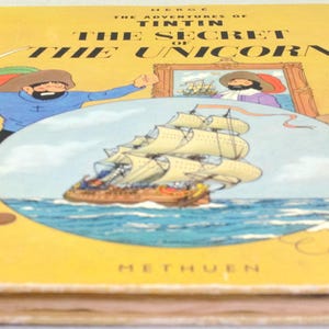May include: A vintage hardcover book titled "The Adventures of Tintin: The Secret of the Unicorn." The cover is yellow with illustrations of Tintin, a ship, and a dog. The book's spine is red, and the title is in bold black letters.