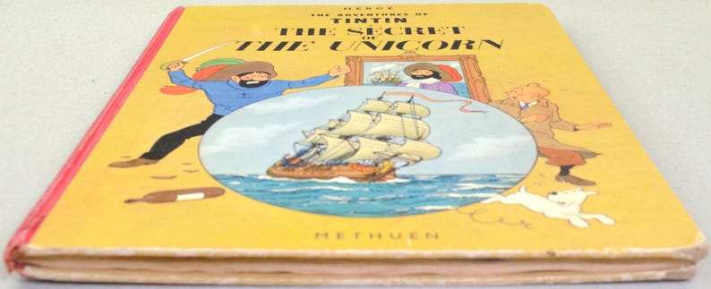 May include: A vintage hardcover book titled "The Adventures of Tintin: The Secret of the Unicorn." The cover is yellow with illustrations of Tintin, a ship, and a dog. The book's spine is red, and the title is in bold black letters.