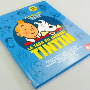 May include: A blue book titled "LA SAGA DU JOURNAL TINTIN" featuring a cartoon image of Tintin and his dog Snowy. The cover also includes the text "DE 1946 &Agrave; 1988" and various cartoon character illustrations.