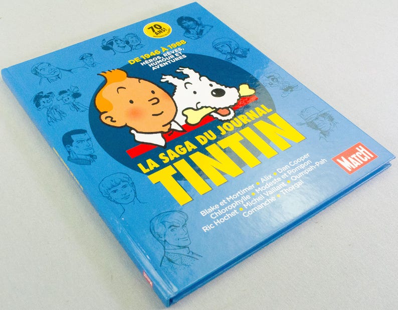 May include: A blue book titled "LA SAGA DU JOURNAL TINTIN" featuring a cartoon image of Tintin and his dog Snowy. The cover also includes the text "DE 1946 &Agrave; 1988" and various cartoon character illustrations.