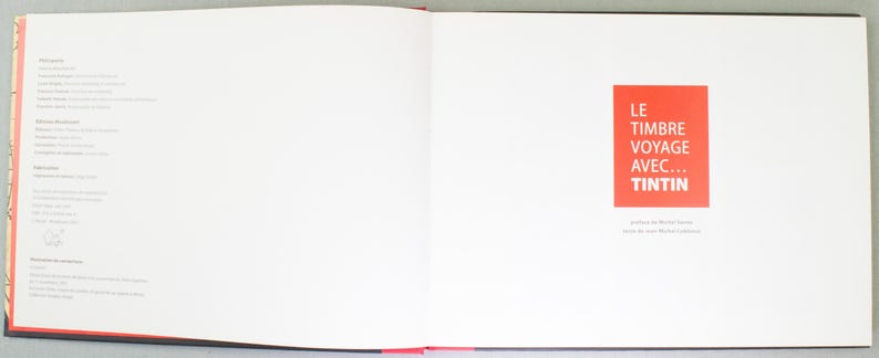 May include: An open book with white pages and red accents. The right page features a red square with white text that reads "LE TIMBRE VOYAGE AVEC... TINTIN". The left page has a list of text in a smaller font.