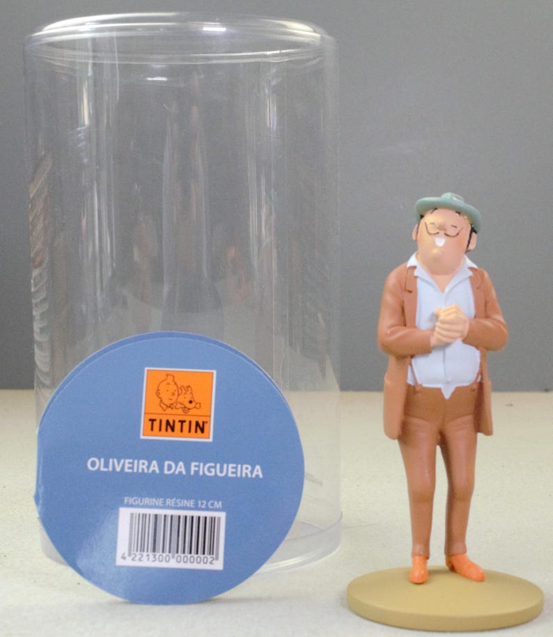May include: A resin figurine of Oliveira da Figueira wearing a tan suit, white shirt, and green hat, standing on a circular base. Next to it is a clear cylindrical display case with a blue circular label featuring the text TINTIN and 12 CM.