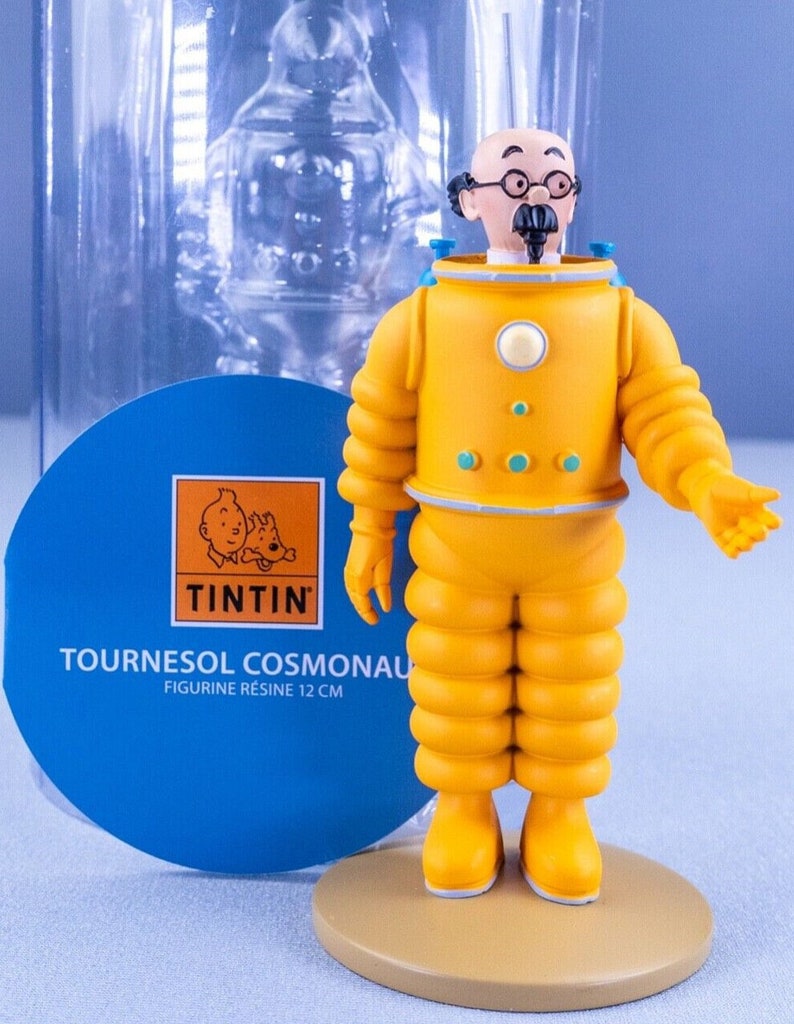 May include: A yellow and white figurine of Professor Calculus from the Tintin comics wearing a yellow space suit. The figurine is 12 cm tall and is made of resin. The figurine is on a tan base and is in a clear plastic box. The box has a blue label with the Tintin logo and the text "Tournesol Cosmonaut Figurine R&eacute;sine 12 cm".