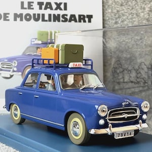 May include: A blue model car with a luggage rack carrying three suitcases. The car has a "TAXI" sign on top and the license plate reads "70V14". The car is parked on a blue base, with a book in the background that reads "LE TAXI DE MOULINSART".