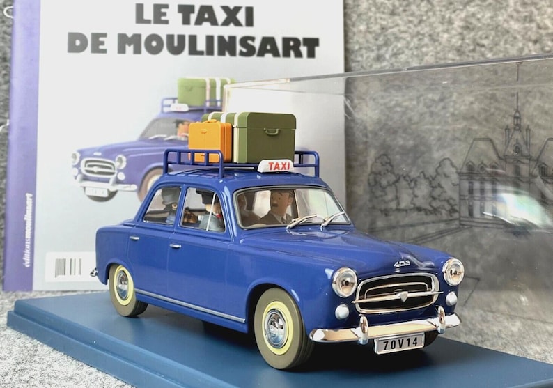 May include: A blue model car with a luggage rack carrying three suitcases. The car has a "TAXI" sign on top and the license plate reads "70V14". The car is parked on a blue base, with a book in the background that reads "LE TAXI DE MOULINSART".