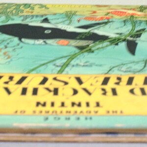 May include: A vintage book titled "Red Rackham's Treasure" by Herg&eacute;. The cover features a black and white shark swimming in a turquoise sea with green seaweed. The title is in black on a yellow banner.