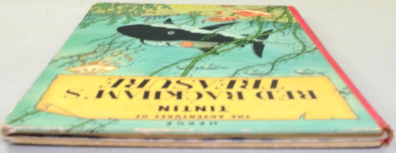 May include: A vintage book titled "Red Rackham's Treasure" by Herg&eacute;. The cover features a black and white shark swimming in a turquoise sea with green seaweed. The title is in black on a yellow banner.
