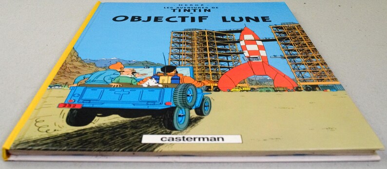 K&ouml;nnte beinhalten: Ein gebundenes Buch mit dem Titel "OBJECTIF LUNE" in franz&ouml;sischer Sprache. Das Cover zeigt einen blauen Jeep mit Passagieren, eine rot-wei&szlig;e Rakete und eine Baustelle. Das Buch hat einen gelben Buchr&uuml;cken und den Namen des Verlags, Casterman.
