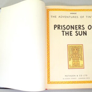 Op de afbeelding: Open boek met de titel "Prisoners of the Sun" uit de serie "De avonturen van Kuifje". De kaft heeft een gele en oranje rand met een illustratie. Uitgegeven door Methuen & Co Ltd.