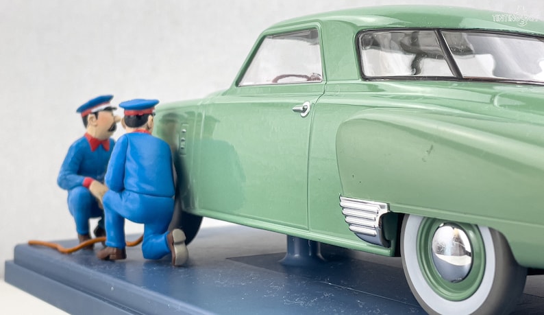 Pu&ograve; includere: Modello di auto d'epoca verde con pneumatici a fascia bianca. Due figure in miniatura in uniforme blu sembrano lavorare sull'auto. L'auto ha dettagli cromati e interni dettagliati, su una base blu.