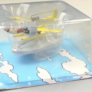 Op de afbeelding: Een geel dubbeldekker model met een pilootfiguur in een doorzichtige plastic display case. Het vliegtuig heeft een propeller en een kleine staart. De verpakking is blauw met witte wolken en tekst.