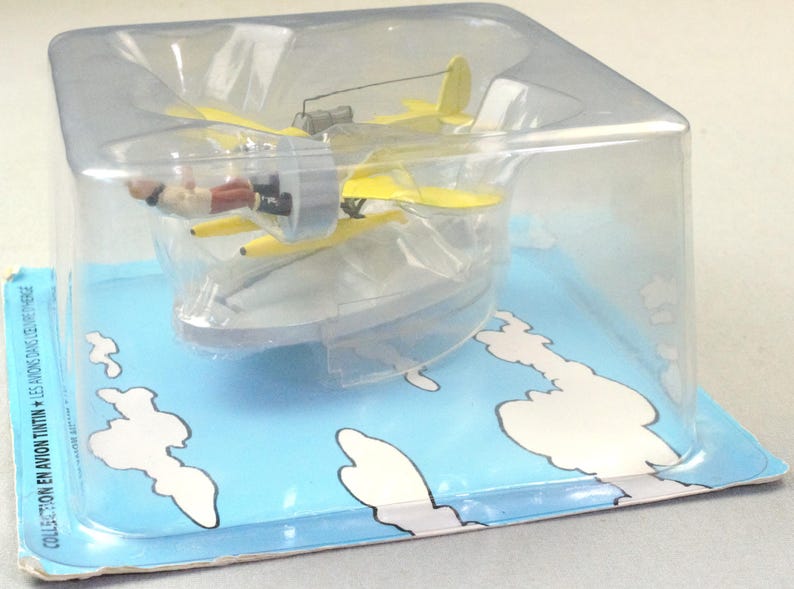 Op de afbeelding: Een geel dubbeldekker model met een pilootfiguur in een doorzichtige plastic display case. Het vliegtuig heeft een propeller en een kleine staart. De verpakking is blauw met witte wolken en tekst.