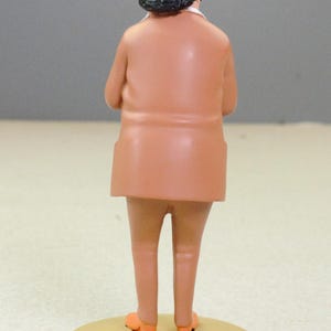 May include: A figurine of a person viewed from behind, wearing a light blue fedora with a black band, a tan suit jacket, and matching tan trousers. The figure stands on a circular tan base, with orange shoes visible at the bottom of the legs.